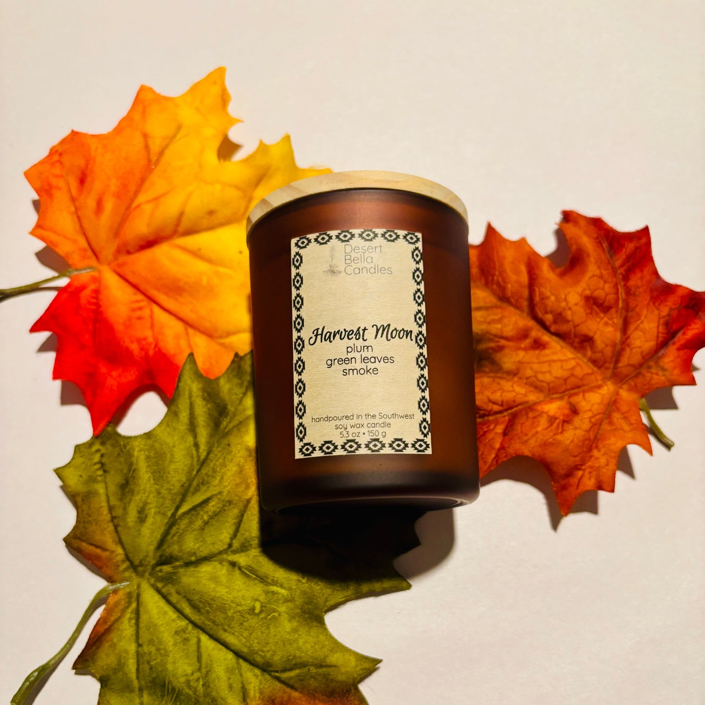 Candle labeled 'Harvest Moon' surrounded by autumn leaves on a neutral background